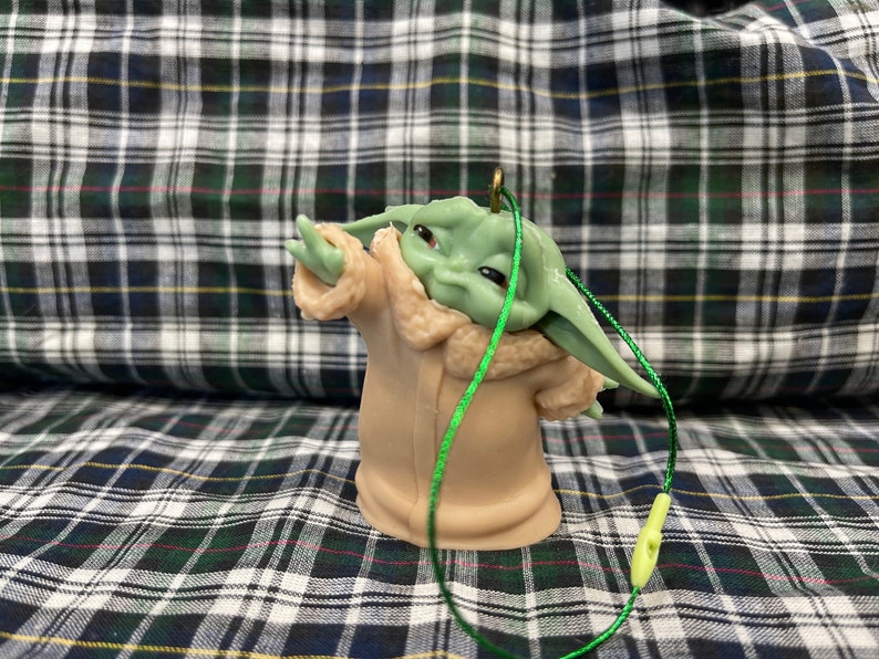 May include: A green and brown plastic bauble featuring a character from the Star Wars franchise, The Mandalorian. The character is wearing a brown coat with a fur trim and has large ears. The bauble has a green string attached to it.