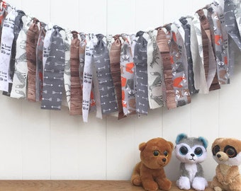 May include: A gray, brown, and white fabric banner with woodland animal and arrow print hangs on a white wall. Three stuffed animals, a bear, a dog, and a monkey, are sitting on a wooden surface in front of the banner.