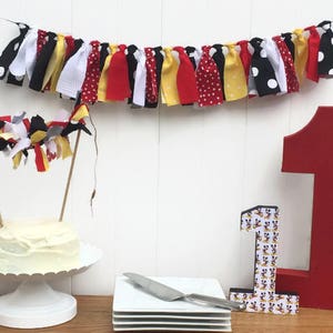 Könnte beinhalten: Ein weißer Kuchen mit rotem und gelbem Frosting ist mit einem Stoffbanner und einer Nummer eins verziert. Das Banner besteht aus roten, gelben, schwarzen und weißen Stoffresten. Die Nummer eins ist aus rotem Holz und mit einem schwarz-weißen Mickey-Mouse-Muster verziert.