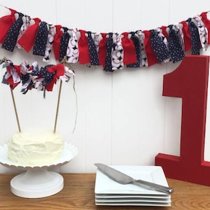 Könnte beinhalten: Ein rot-weiß-blaues Stoffbanner mit Baseball-Motiven hängt über einem weißen Kuchenständer mit einem weiß glasierten Kuchen. Zwei Stoffbanner sind mit Holzstäbchen am Kuchen befestigt. Eine große rote Nummer eins steht auf einem Holztisch.
