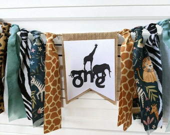 May include: A burlap banner with the word "one" in black letters and a silhouette of a giraffe, elephant, and zebra. The banner is decorated with fabric strips in various animal prints, including zebra, giraffe, and jungle foliage.