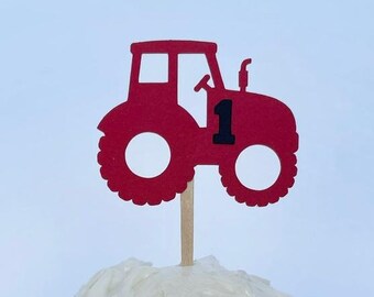 May include: Red tractor cake topper with the number 1 on the side. The tractor is made of paper and is attached to a wooden stick.