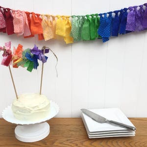Könnte beinhalten: Ein kleiner weißer Kuchen auf einem weißen Kuchenständer, verziert mit einem Regenbogen-Topper. Ein Regenbogen-Stoffbanner hängt darüber. Ein Stapel weißer quadratischer Teller und ein Messer liegen auf einem Holztisch.