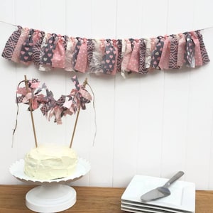 Könnte beinhalten: Ein weißer Kuchen mit rosa Zuckerguss ist mit einer rosa und grauen Stoffgirlande verziert. Der Kuchen steht auf einem weißen Kuchenständer. Auf einem Holztisch liegen vier weiße Teller übereinander.