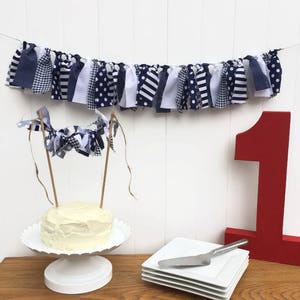 Könnte beinhalten: Ein weißer Kuchen mit weißem Frosting steht auf einem weißen Kuchenständer. Über dem Kuchen hängt ein blaues und weißes Stoffbanner. Das Banner besteht aus verschiedenen Stoffmustern. Eine rote Nummer eins steht rechts neben dem Kuchen.
