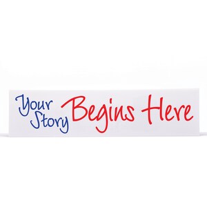 6 x 24 inch real estate sign rider against white backdrop. It is made of aluminum metal and has a white background with bold red and blue scripted font. Says your story begins here.