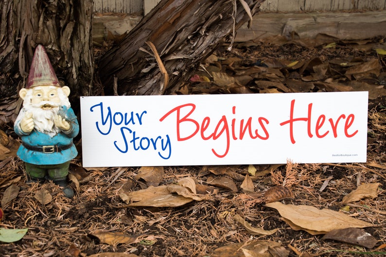 6 x 24 inch real estate rider against fall leaves backdrop. It is made of aluminum metal and has a white background with bold red and blue scripted font. Says your story begins here. Rider is next to a  9 inch gnome statue.