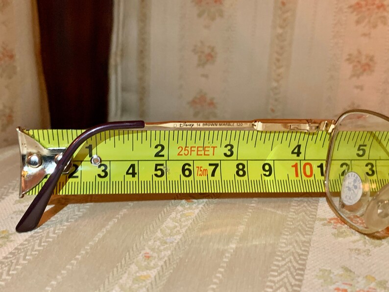 May include: Gold-tone eyeglasses with a purple arm, measured with a yellow tape measure. The glasses are marked "Disney 14 BROWN MARBLE 120". The tape measure shows a length of approximately 4 inches.