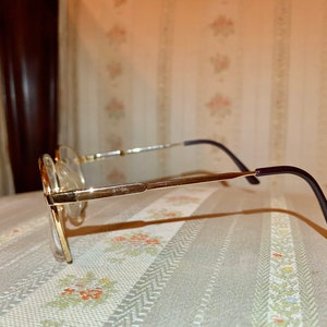 May include: Gold-rimmed eyeglasses with clear lenses. The frames have a rectangular shape with a thin, gold metal frame and dark purple temples. The glasses are set against a patterned background.