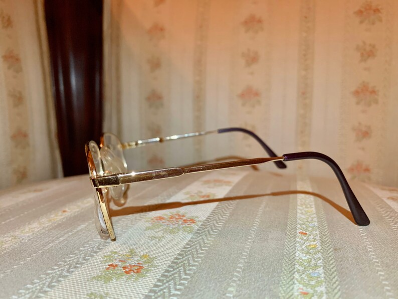 May include: Gold-rimmed eyeglasses with clear lenses. The frames have a rectangular shape with a thin, gold metal frame and dark purple temples. The glasses are set against a patterned background.
