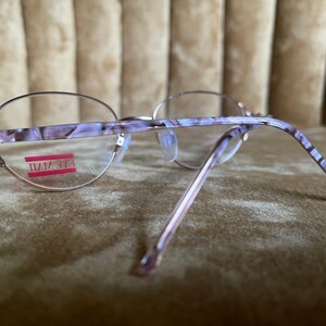 May include: A pair of oval-shaped eyeglasses with a pink and white marbled frame. The frame has a silver metal accent on the side. The eyeglasses are labeled "EYE MAIL" on the right lens.