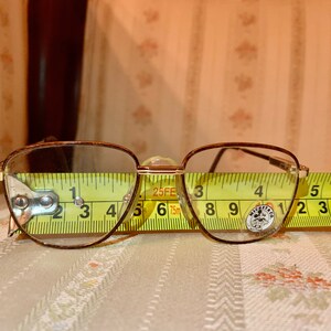May include: Vintage eyeglasses with a brown metal frame and clear lenses. The glasses are measured with a yellow tape measure, showing a width of approximately 5 inches. The lenses have a rounded rectangular shape.