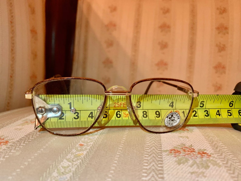 May include: Vintage eyeglasses with a brown metal frame and clear lenses. The glasses are measured with a yellow tape measure, showing a width of approximately 5 inches. The lenses have a rounded rectangular shape.