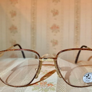 May include: Gold-toned eyeglasses with a rectangular frame and clear lenses. The frames have a textured pattern and black temples. A small sticker with the text "Mickey & Co" and a Disney logo is visible on the right lens.