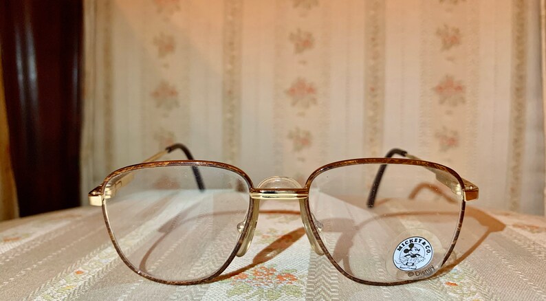 May include: Gold-toned eyeglasses with a rectangular frame and clear lenses. The frames have a textured pattern and black temples. A small sticker with the text "Mickey & Co" and a Disney logo is visible on the right lens.