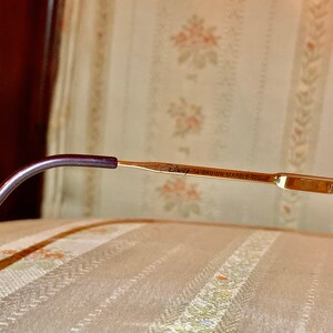 May include: Close-up of eyeglasses with a gold-colored frame and a dark purple temple. The text "Disney 14 BROWN MARBLE 120" is printed on the frame. The glasses are resting on a patterned surface.