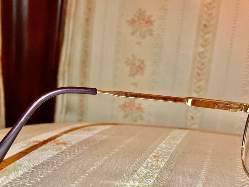 May include: Close-up of eyeglasses with a gold-colored frame and a dark purple temple. The text "Disney 14 BROWN MARBLE 120" is printed on the frame. The glasses are resting on a patterned surface.