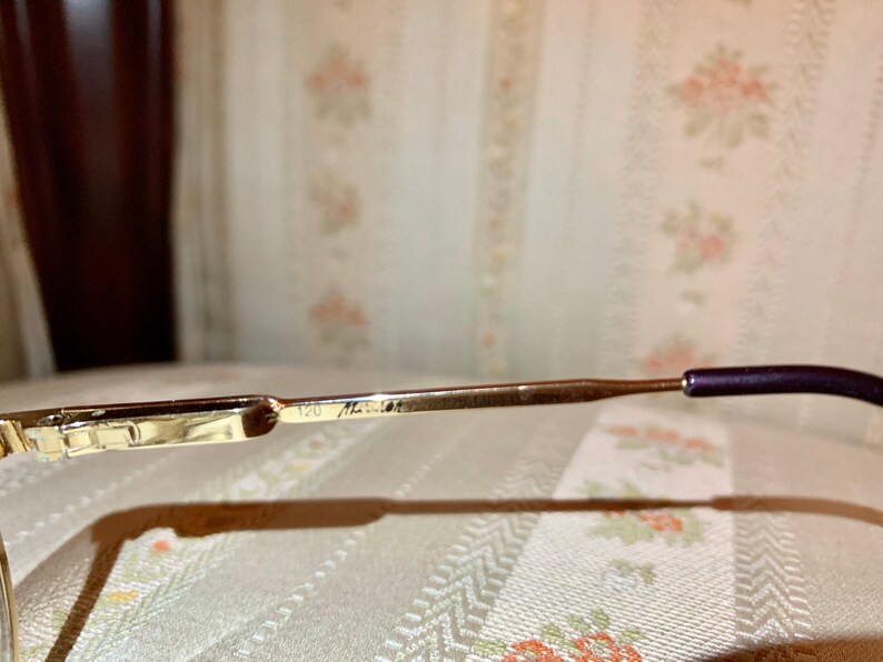 May include: Close-up of a pair of eyeglasses. The frames are gold-colored with a rectangular design on the front. The temple arms are a gradient from gold to brown, ending with a dark purple earpiece. The number "120" and other text are visible on the frame.