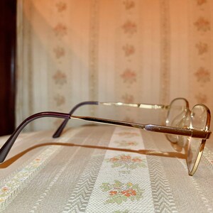 May include: A pair of eyeglasses with a gold-colored metal frame and clear lenses. The temples are a dark purple color. The glasses are resting on a patterned surface.