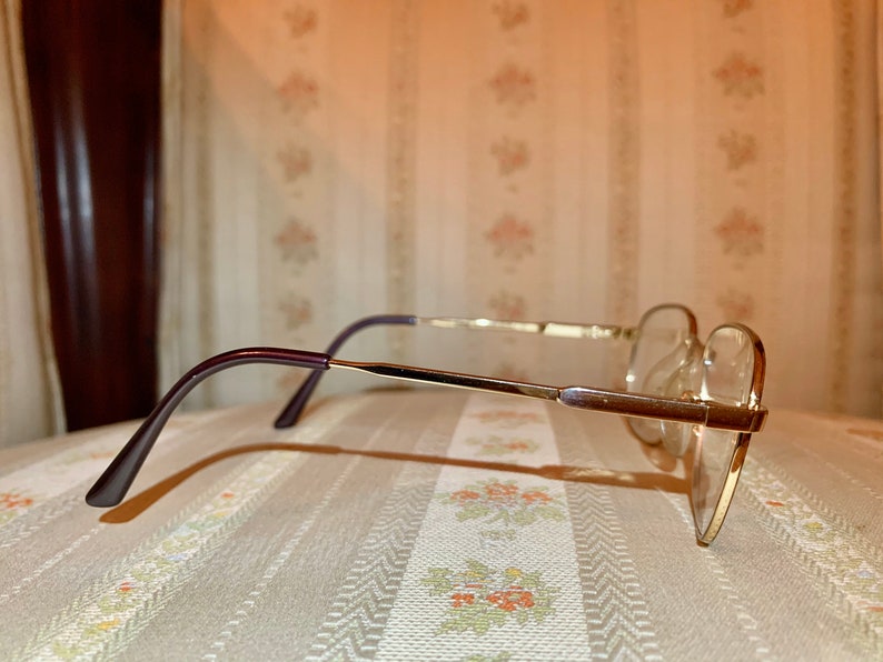 May include: A pair of eyeglasses with a gold-colored metal frame and clear lenses. The temples are a dark purple color. The glasses are resting on a patterned surface.