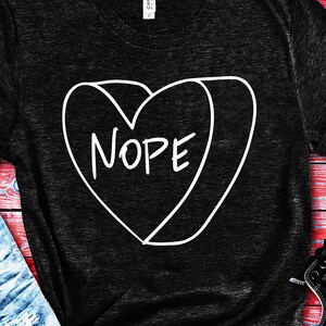 Puede incluir: Una camiseta negra con un contorno blanco de un corazón que dice "NOPE" en su interior.