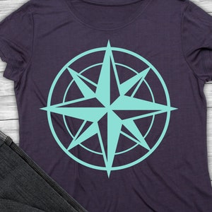 Könnte beinhalten: Ein dunkelviolettes T-Shirt mit einem türkisfarbenen Kompass-Design. Der Kompass hat eine Sternform mit acht Zacken und ist von einem Kreis umgeben.