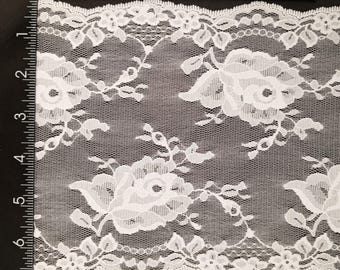 May include: White floral lace fabric featuring a repeating pattern of large roses and smaller flowers. The lace has a scalloped edge and is measured in centimetres. Ideal for craft projects.