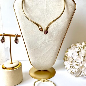 Puede incluir: Un conjunto de collar gargantilla, pulsera y pendientes de tono dorado con dos acentos de cristal rosa en cada pieza.