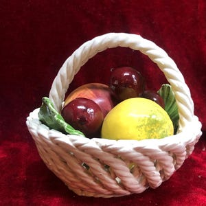 May include: A white ceramic basket with a braided handle and woven sides, filled with a collection of glossy, hand-painted ceramic fruits including a bright yellow lemon, dark red cherries, and a multi-coloured peach with green leaves.