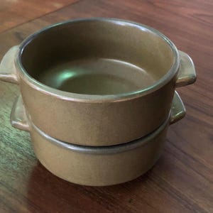 May include: Two stacked round ceramic bowls with a matte brown finish. Each bowl features two small, integrated side handles and a smooth, circular rim. The bowls have a consistent earthy tone and a simple, functional design for serving food.