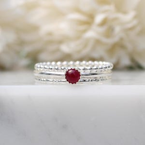 Op de afbeelding: Een stapelbare ringenset bestaande uit drie zilveren banden. De bovenste en onderste banden hebben een kraaltextuur, terwijl de middelste band glad is en een kleine, ronde, dieprode edelsteen bevat in een geschulpte zilveren zetting.