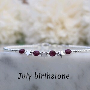 Può includere: Un delicato bracciale a catena in argento con piccole perle rotonde di rubino sfaccettato rosa scuro che si alternano a ciondoli a forma di stella in argento lucido. Il bracciale è mostrato sopra il testo July birthstone in carattere serif.