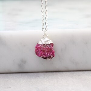 May include: A silver chain necklace featuring a raw ruby pendant. The ruby is a deep pink-red hue with a textured surface. The top of the ruby is capped with silver. The necklace is displayed on a white marble surface.