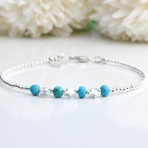 Op de afbeelding: Een delicate zilveren armband met kleine, ronde kralen en turquoise gekleurde kralen. De armband heeft ook kleine stervormige bedels. De sluiting is zichtbaar aan het einde van de armband.