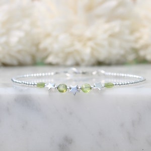 May include: A silver bracelet with small, round beads, star-shaped charms, and light green gemstones. The bracelet is presented on a white marble surface, with a soft, blurred floral backdrop. The bracelet is approximately 18 cm in length.