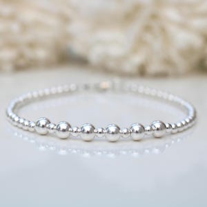 Könnte beinhalten: Ein filigranes Silberarmband mit kleinen, runden Perlen. Das Armband hat ein schlichtes Design mit größeren, polierten Perlen, die zwischen den kleineren angeordnet sind. Der Schmuck wird auf einer weißen Oberfläche mit einem weichen, verschwommenen Hintergrund präsentiert.