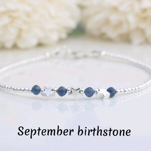 May include: A delicate silver bracelet featuring small silver beads and accents of blue gemstones and star-shaped charms. The text "September birthstone" is displayed below the bracelet. This bracelet is a piece of jewelry.