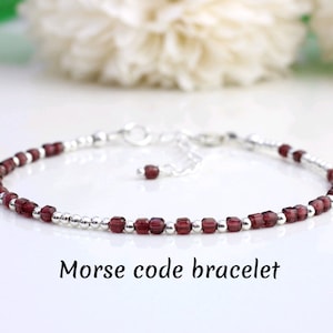 Pode incluir: Uma delicada pulseira de prata com pequenas contas vermelhas escuras e detalhes prateados. A pulseira está rotulada como "Morse code bracelet" em texto preto. A pulseira é exibida em uma superfície branca com flores brancas desfocadas ao fundo.