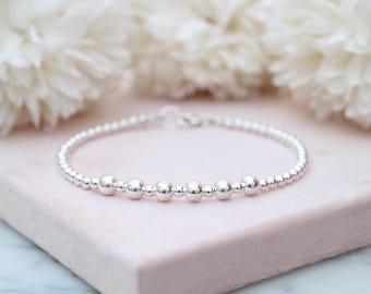 Könnte beinhalten: Ein Armband aus silberfarbenen Perlen, das eine Reihe kleiner, gleichmäßiger Metallkugeln mit etwas größeren, polierten Silberperlen kombiniert, die in einem sich wiederholenden Muster entlang des kreisförmigen Schmuckstücks angeordnet sind.