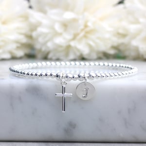 May include: Silver beaded bracelet with a cross charm and a personalised initial charm. The initial charm is a silver disc with the letter "J" engraved on it.