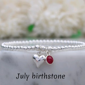 Può includere: Un braccialetto elastico di perline argentate con un ciondolo a forma di cuore in argento lucido e un piccolo ciondolo circolare rosso che rappresenta la pietra portafortuna di luglio. Il testo July birthstone è stampato in nero.