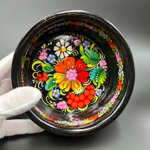 Puede incluir: Un pequeño cuenco redondo de madera negra con un vibrante diseño floral. El interior presenta una disposición central de flores coloridas en rojo, naranja, amarillo y blanco, rodeadas de hojas verdes y flores azules y rosas más pequeñas. El borde es negro con un acento dorado.