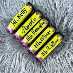 May include: Five yellow glitter pencil-shaped items with black text. The pencils have black and gold glitter tips, pink erasers, and silver bands. The text includes names like "Mr. Kelly" and "Apple".