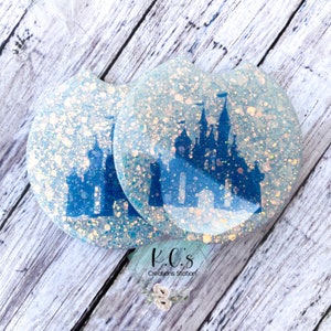 May include: Two round, white, glitter-filled coasters with a blue silhouette of a castle in the center. The coasters are on a light wood surface. The text "K.C.'s Creations Station" is visible below the coasters.