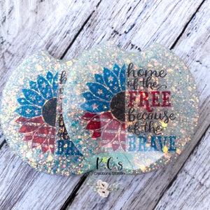 May include: Two round white coasters with a glittery finish. Each coaster features a red, white, and blue sunflower design. The text "home of the FREE because of the BRAVE" is printed on one coaster. The text "K.C.'s Creations Station" is printed on the bottom of the coaster.