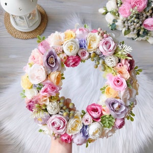 May include: A pastel pink, white, and yellow floral wreath with greenery and small white balls. The wreath is made of artificial flowers and is perfect for a spring or summer wedding.