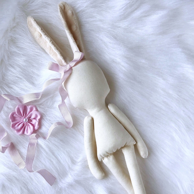 May include: A white fabric bunny doll with long ears and a pink ribbon around its head. The doll is lying on a white fluffy surface.