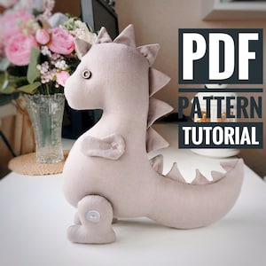 Może przedstawiać: Pluszowa zabawka w kształcie dinozaura w jasnobrązowym kolorze z guzikowym okiem i guzikiem na stopie. Zabawka leży na białym tle z napisem "PDF PATTERN TUTORIAL" w kolorze czarnym.