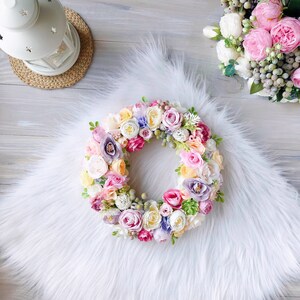 May include: A pastel-colored floral wreath with pink, white, yellow, and purple flowers. The wreath is made of silk flowers and is perfect for decorating a home or office.