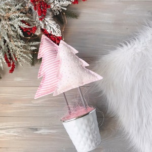 May include: Two pink fabric Christmas trees in a white metal pot with pink and white filler. The trees are decorated with white polka dots and pink stripes.